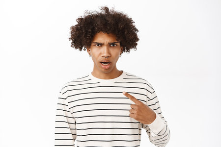 Shocked, offended young hispanic boy, pointing finger at himself, staring upset at camera, standing over white backgroundの写真素材