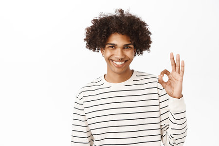 Close up portrait of smiling hispanic guy shows okay, ok sign, approves smth, recommends, stands over white studio backgroundの写真素材