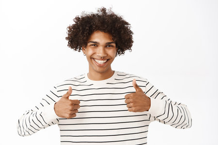 Close up portrait of smiling handsome hispanic guy, showing thumbs up, pleased by smth, recommending course or product, standing over white studio backgroundの写真素材