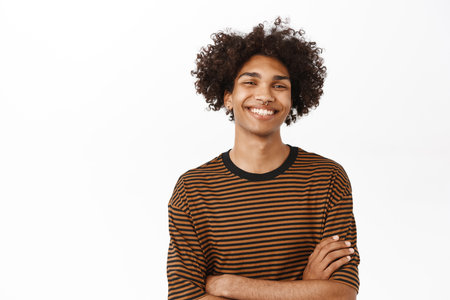 Close up of handsome modern hispanic guy with afro hair, smiling happy, cross arms on chest and looking confident, standing over white backgroundの写真素材