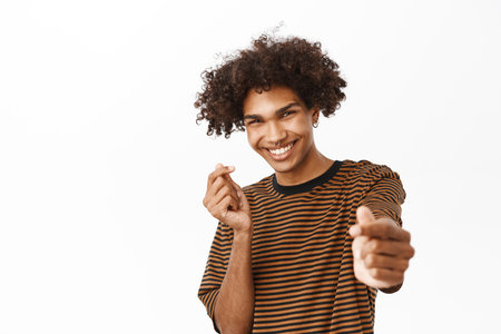 Close up of handsome curly guy shows finger hearts and smiling cute at camera, standing over white backgroundの写真素材