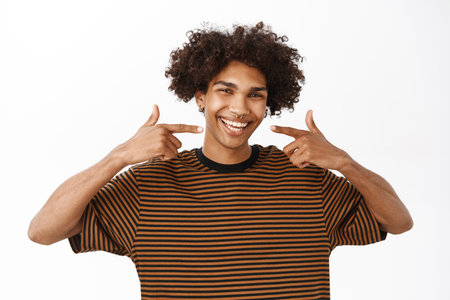 Happy handsome boy with curly hair, pointing at his white teeth, showing healthy perfect smile, stands over studio backgroundの写真素材