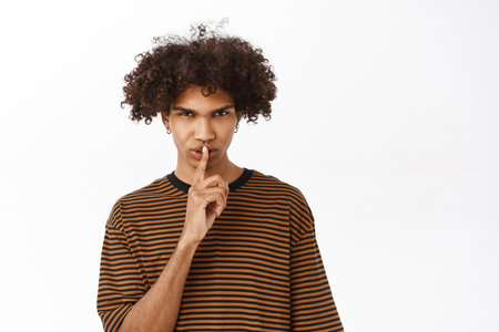 Keep quiet. Serious hispanic boy shushing, say shh, showing taboo gesture with finger pressed to his mouth, standing over white studio backgroundの写真素材