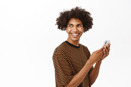 Portrait of young modern guy using mobile phone, looking behind his shoulder and smiling, standing with smartphone against white backgroundの写真素材