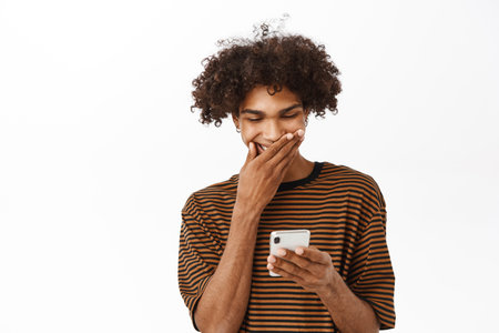 Portrait of young man reading on mobile phone and laughing, watching smth funny on smarpthone app, standing over white backgroundの写真素材