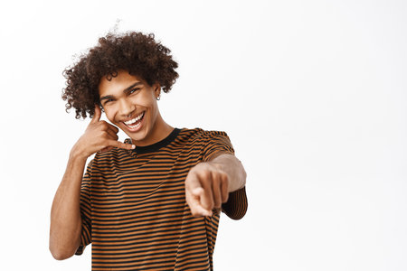 Call me. Handsome hispanic guy with curly hair, showing mobile phone near ear and pointing finger at camera, inviting you, standing over white backgroundの写真素材