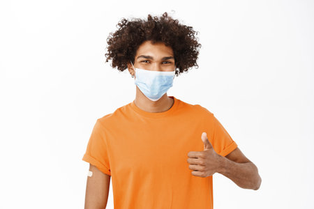 Young hispanic man recommends do covid 19 vaccine shot, join vaccination campaign, showing thumbs up, wearing medical face mask, standing over white backgroundの写真素材
