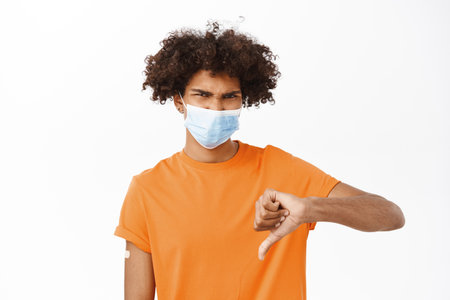 Disappointed young man in medical face mask, showing thumbs down, he has patch on arm after covid-19 vaccination, dislike antivaxxers, standing over white backgroundの写真素材