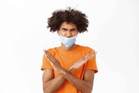 Man showing stop sign, wearing medical face mask wrong, showing thumbs down, he has patch on arm after covid-19 vaccination, dislike smth, standing over white backgroundの写真素材