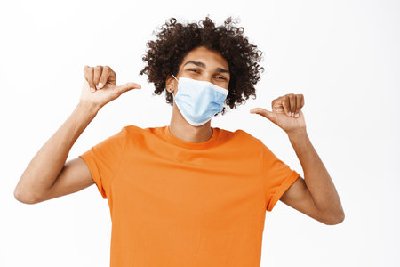 Image of happy young handsome guy pointing at his medical face mask, asking to wear it from covid-19, standing over white backgroundの写真素材
