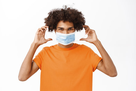 Image of handsome young man smiling, showing how to wear medical face mask during covid-19 pandemic, standing in orange tshirt over white backgroundの写真素材