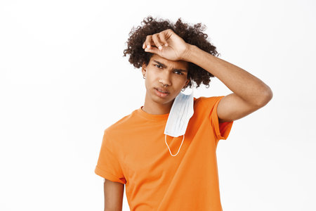 Image of tired hispanic guy takes off medical face mask and wipes sweat off forehead, looking ill, standing over white studio backgroundの写真素材