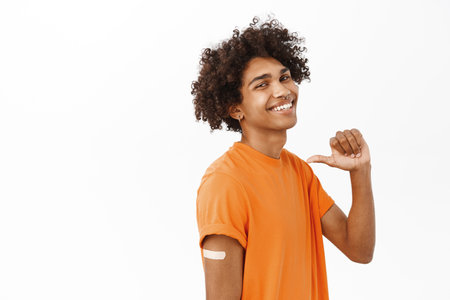 Handsome smiling young man, did covid-19 vaccination shot on arm, showing his patch, pointing at himself, standing over white backgroundの写真素材