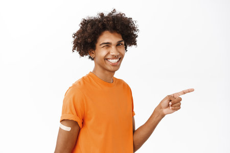 Covid-19 vaccination campaign. Smiling handsome boy with coronavirus vaccine shot, pointing right at logo or banner, wearing orange tshirt, white backgroundの写真素材
