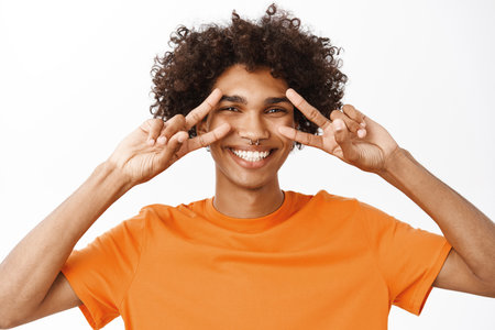 Close up portrait of smiling hispanic man showing peace, v-sign near eyes and looking happy, positive people and emotions, standing over white backgroundの写真素材