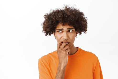 Image of young hispanic guy feeling anxious, worried by smth, biting fingers and lookign concerned, standing over white backgroundの写真素材