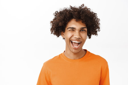 Close up of handsome hispanic man with curly hair, smiling and laughing, looking sassy, wearing orange t-shirt, standing over white studio backgroundの写真素材