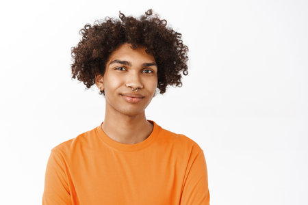 Close up of handsome curly boy with piercing, queer guy in orange t-shirt smiling, standing over white backgroundの写真素材