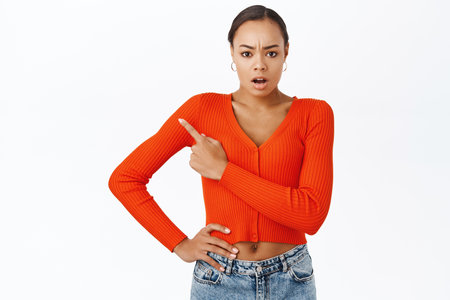 Shocked and concerned african american woman, pointing finger at upper left corner, looking with worried face, standing over white backgroundの写真素材