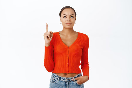 Disappointed young latina woman, pointing finger up, looking with skeptical, upset face expression, standing over white backgroundの写真素材