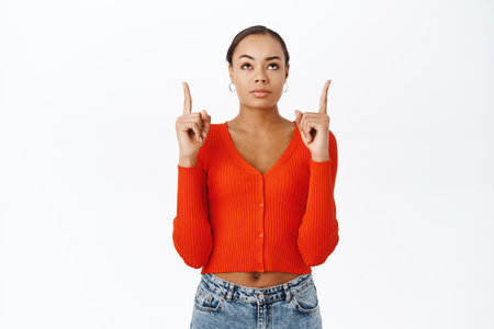 Portrait of young woman looks with doubt and skepticism, pointing fingers up at logo, promo offer, stands over white backgroundの写真素材