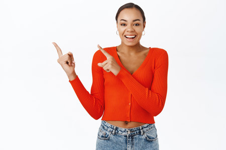 Enthusiastic young latina woman, pointing fingers at upper left corner, smiling and laughing, showing advertisement, promo banner, standing over white backgroundの写真素材