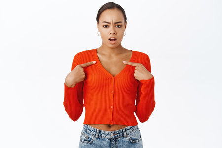 Shocked, offended latina woman pointing at herself, looks insulted and upset, stands over white studio backgroundの写真素材