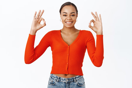 Excellent, very good. Smiling young latina woman, shows okay, praise gesture, recommends smth, stands over white backgroundの写真素材