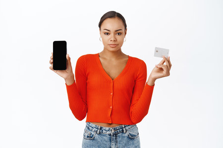 Serious looking 30 years old woman showing mobile phone screen and credit card, frowning with upset face, standing over white backgroundの写真素材