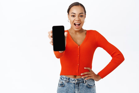 Portrait of 30 years old woman showing smartphone screen, demonstrates app interface, webstore, stands with mobile phone against white backgroundの写真素材