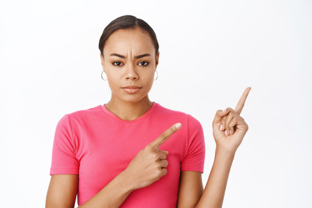 Close up of upset black woman, pointing fingers right, showing advertisement with disappointed face, standing over white backgroundの写真素材
