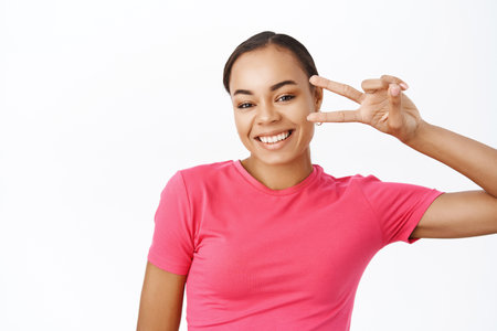 Close up of smiling black female model, shows peace, v-sign near eye and looks happy, wears pink tshirt, stands over white backgroundの写真素材