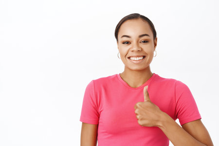 Close up of enthusiastic girl shows her support, likes smth, thumbs up in approval, stands over white studio backgroundの写真素材