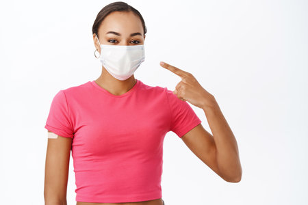 Portrait of girl with patch from covid-19 vaccine, pointing at her medical face mask, standing over white backgroundの写真素材