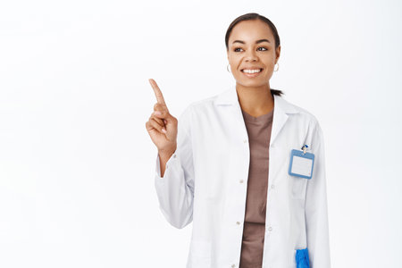 Woman doctor in white coat, pointing finger and looking at banner, showing medical clinic logo on copy space, standing over white backgroundの写真素材