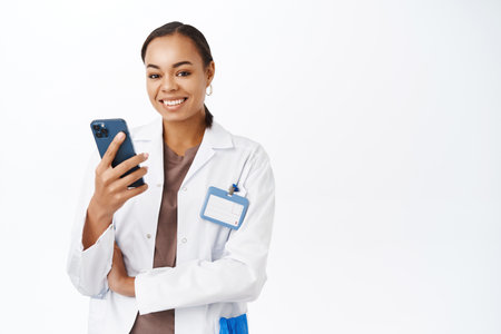 Online medical help. Young female doctor, therapist holding mobile phone, answer patients messages on healthcare app, standing over white backgroundの写真素材