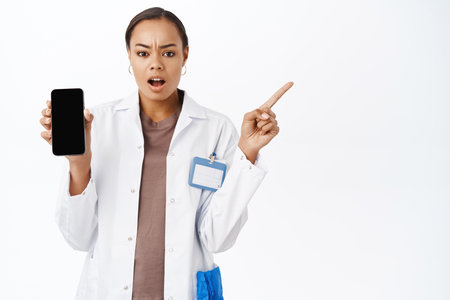 Shocked woman therapist, showing mobile phone screen, pointing right at banner, looking concerned, standing over white backgroundの写真素材