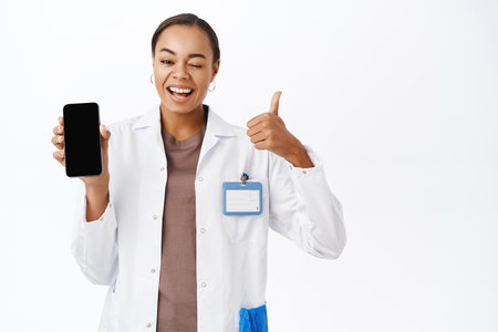 Woman doctor shows thumbs up and smartphone screen app, mobile healthcare application interface, standing over white backgroundの写真素材