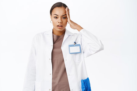 Troubled female doctor, holds hand on head, look concerned and complicated, stands against white backgroundの写真素材