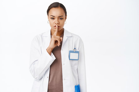 Woman doctor shushing, showing taboo sign, keep quiet gesture, standing over white studio backgroundの写真素材