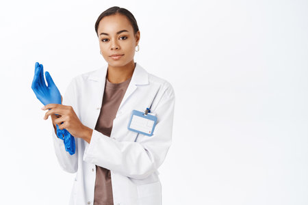 Doctor puts on gloves, prepare for patient checkup, smiling enthusiastic at camera, standing over white backgroundの写真素材