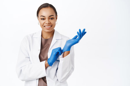 Female therapist, doctor puts on medical gloves, ready to see patient, take samples at laboratory, smiling happy at camera, white backgroundの写真素材
