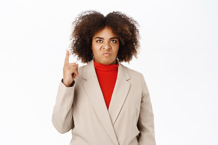 Angry businesswoman looks up, points at top with jealous, annoyed face expression, stands over white backgroundの写真素材