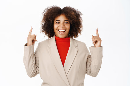 Enthusiastic black woman, pointing fingers up, showing logo or company banner on top, standing in suit over white backgroundの写真素材
