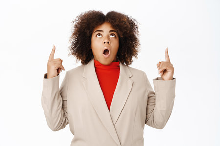 Portrait of surprised black woman, saleswoman pointing fingers up, looks impressed and amazed by company banner, standing over white backgroundの写真素材