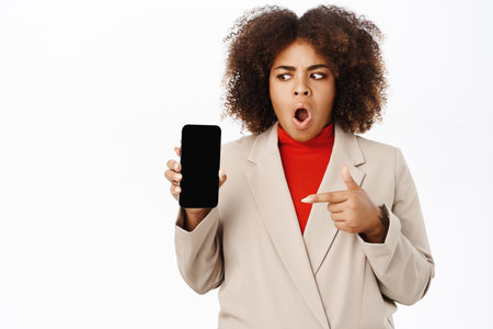 Technology. Shocked african american businesswoman, points and looking at smartphone screen with startled face expression, stands over white backgroundの写真素材