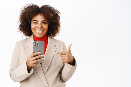 Portrait of smiling african american woman records video, takes photo on smartphone and points at mobile phone camera, stands over white backgroundの写真素材