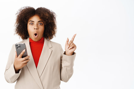 Amazed black businesswoman, holds smartphone and points aside at banner or logo, stands over white studio backgroundの写真素材