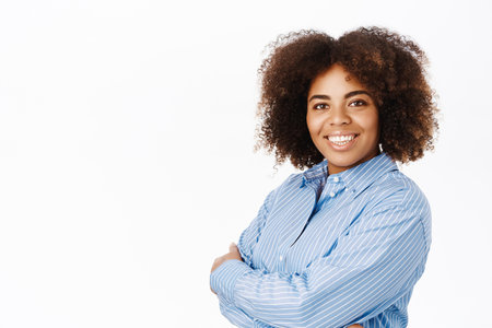 Portrait of young ambitious african american woman, cross arms on chest, smiles and looks confident, stands over white backgroundの写真素材