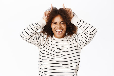 Funny girl shows finger horns on head and laughs, looking joyful, standing over white studio backgroundの写真素材